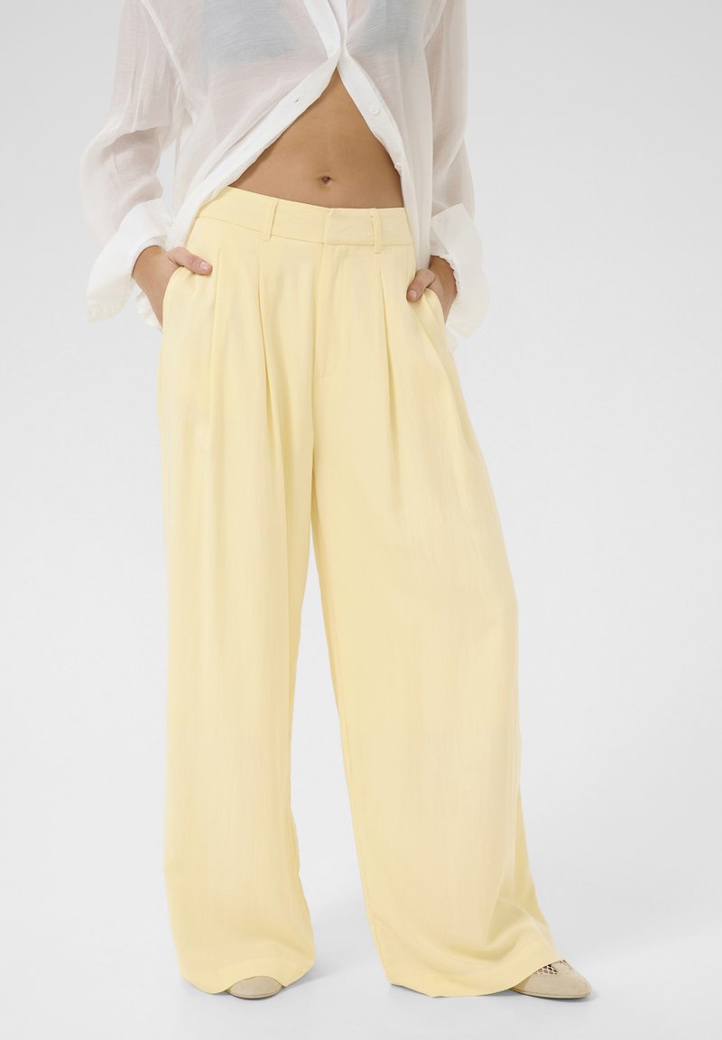Pantaloni larghi a plissettatura gialla con vita alta e tessuto liscio, abbinati a una camicia bianca trasparente con bottoni.