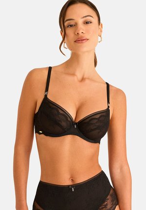 Femme portant un soutien-gorge en dentelle noire et une culotte taille haute en dentelle, posant devant un fond clair uni.