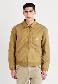 Giacca in cotone marrone con chiusura a zip frontale, colletto e tasca con patta. Presenta un taglio relax e dettagli di cucitura visibili.