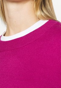 Fuchsia gestrickter Pullover mit weißem Rundhalsausschnitt. Der Stoff wirkt weich mit einer glatten Textur und subtilem Glanz. Nahaufnahme des Halsbereichs.