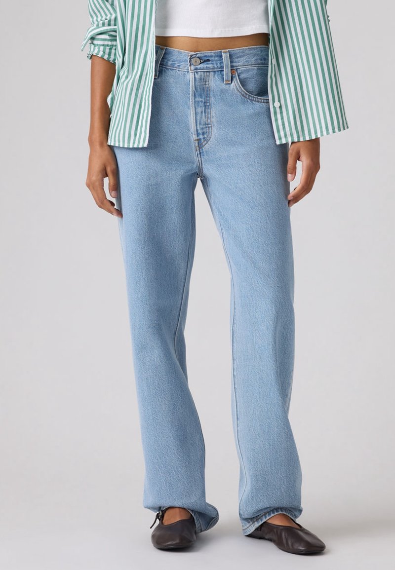 Personne portant un jean droit bleu clair, des ballerines noires, un top court blanc et une chemise rayée verte et blanche avec les manches retroussées.