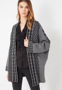 Mujer con cabello largo vistiendo un cárdigan de punto gris con patrones geométricos negros sobre una blusa negra con volantes y pantalones negros.