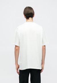 Biała bawełniana koszulka z krótkim rękawem, okrągłym dekoltem i luźnym krojem. Ma gładką teksturę i prosty wzór, bez widocznych akcentów.