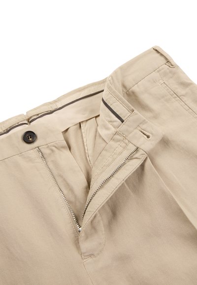 Pantalon beige avec braguette ouverte, fermeture par bouton, passants de ceinture et couture intérieure visible de la ceinture sur fond blanc.