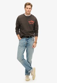 Sudadera gris oscura con gráfico rojo, jeans de corte relajado en azul claro, zapatillas beige y un cinturón negro, de pie sobre un fondo blanco.