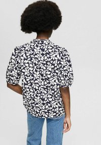 Blouse fleurie avec un fond bleu marine, motif de feuilles blanches, manches courtes bouffantes et encolure froncée ; associée à un jean bleu.