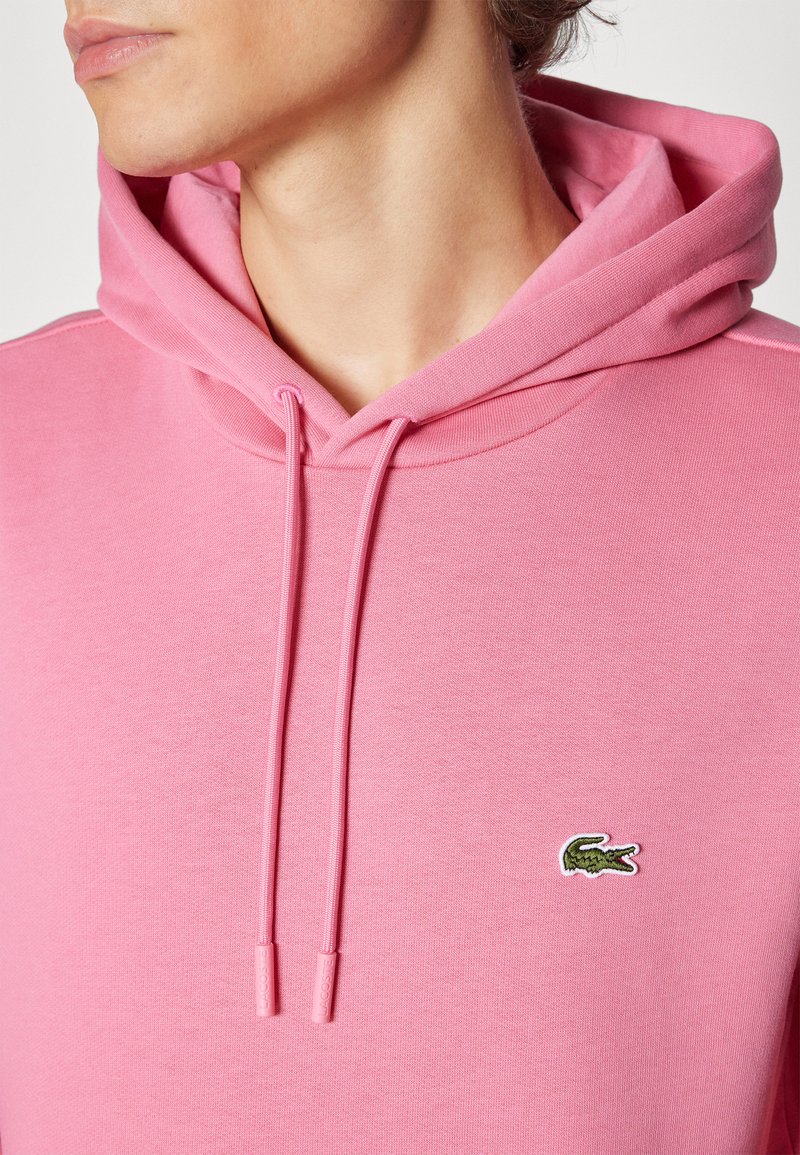 Hoodie rose en tissu doux, avec une poche kangourou, des cordons de serrage et un petit logo de crocodile brodé en vert sur la poitrine.
