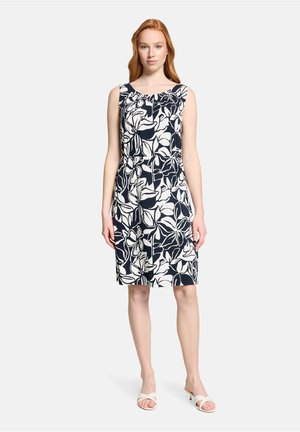 Kobieta stojąca w sukience bez rękawów o długości do kolan z dużym czarno-białym kwiatowym wzorem oraz białych sandałach na obcasie na jednolitym tle.