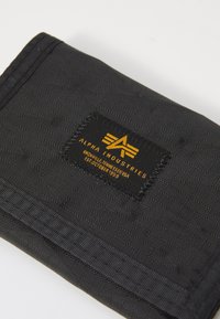 Black fabric wallet with a textured surface, featuring a sewn-on black label with gold lettering and a logo. Simple, compact design.