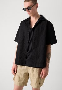 Camicia nera a maniche corte con un motivo testurizzato, indossata sopra una canotta nera e abbinata a pantaloni corti in lino beige.