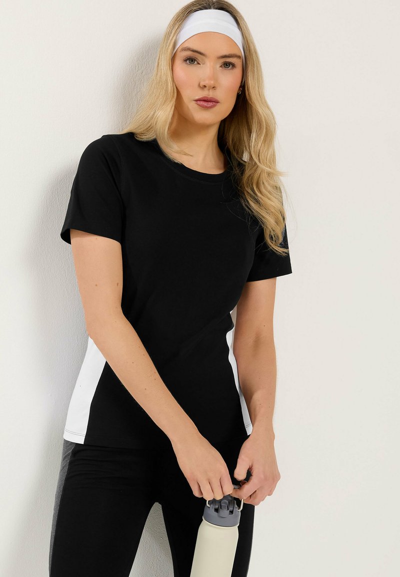 Mujer con conjunto deportivo en blanco y negro sosteniendo una botella de agua, con una cinta blanca en la cabeza, de pie frente a una pared lisa y clara.