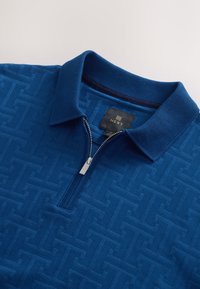 Blauw poloshirt met een gestructureerd patroon, korte kraag en halve rits. Het materiaal is lichtgewicht en heeft een subtiel geometrisch design.