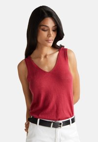 Top sin mangas rojo con un diseño de escote en V y textura suave, combinado con pantalones blancos y un cinturón negro. Silueta simple y moderna.