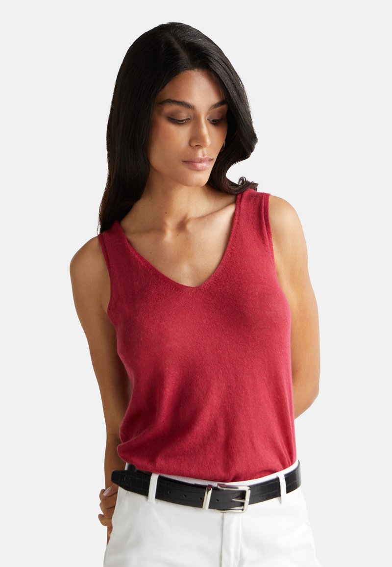 Top sin mangas rojo con un diseño de escote en V y textura suave, combinado con pantalones blancos y un cinturón negro. Silueta simple y moderna.