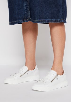 Persoon draagt witte lage sneakers met ritssluitingen aan de zijkant en blauwe denim kniebroek, staand op een effen oppervlak.