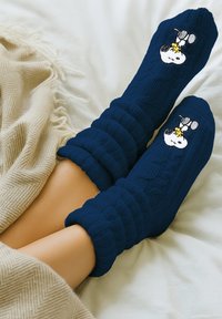Calzini lavorati a maglia blu navy con polsini a coste presentano un design di Snoopy e Woodstock. Circondati da una coperta beige, esaltando la texture e il contrasto di colore.
