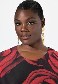 Primo piano di una persona che indossa un top nero con grandi motivi di rose rosse, con orecchini rotondi d'oro e pelle liscia con trucco naturale.