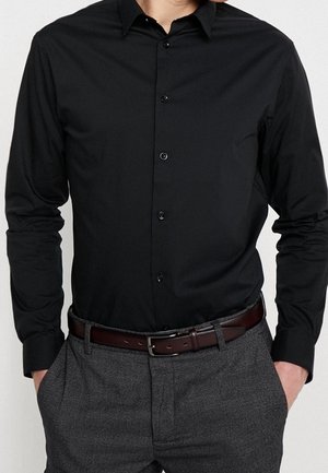 Homme portant une chemise noire cintrée boutonnée rentrée dans un pantalon gris avec une ceinture en cuir marron, mains dans les poches, torse visible.