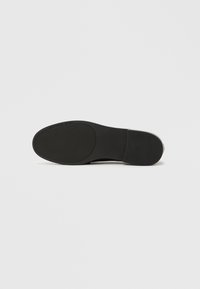 Μαύρη σόλα επίπεδου παπουτσιού με υφασμένη επιφάνεια από καουτσούκ, ελαφριά καμπύλη στη μύτη, ανάγλυφο λογότυπο και χωρίς ορατή φτέρνα.