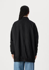 Schwarze leichte Jacke mit hohem Kragen, elastischem Saum und Bündchen. Sie hat eine glatte Textur und eine lockere Passform, kombiniert mit blauen Jeans.