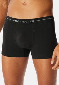 Mandlig model iført sorte Schiesser boxershorts, som viser fronten samt delvist torso og lår mod en hvid baggrund.