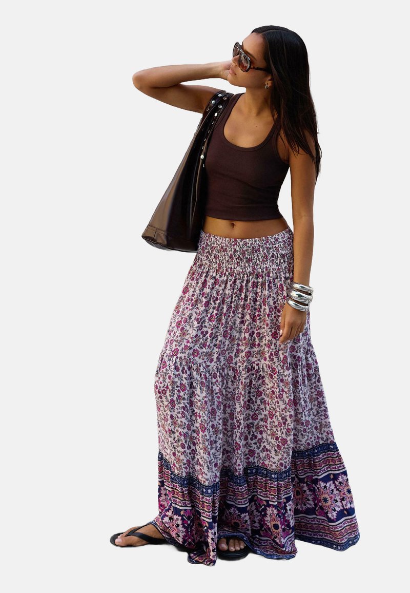 Maxi-Rock mit Blumenmuster und gerafftem Bund, versehen mit pinken und lila Mustern und einem kontrastierenden navyblauen unteren Saum. Kombiniert mit einem dunklen Tanktop und Accessoires.