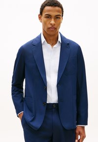 Νεαρός άνδρας που φορούσε κοστούμι σε μπλε χρώμα ραμμένο στα μέτρα του και λευκό πουκάμισο, στέκεται με τα χέρια στις τσέπες μπροστά σε απλό λευκό φόντο.