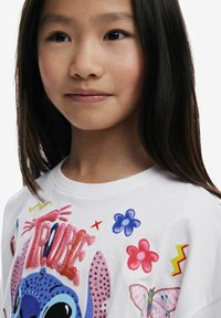 Una camiseta blanca de algodón presenta gráficos de dibujos animados coloridos que incluyen un personaje azul, flores y una mariposa, con acentos brillantes y patrones.
