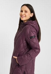 Paarse gewatteerde jas met capuchon, voorzien van diagonale stiksels, zijzakken en een logopatch op de mouw. Glad, glanzend materiaal.