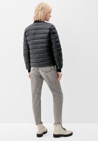 Crna puffer jakna s horizontalnim šavovima, sivi traperice s razderanim rubovima i krem čizme s crnim potplatima. Model je okrenut leđima.
