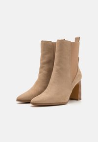 Bottines beiges en suède, avec un bout pointu et un talon épais aux rayures horizontales. Panneaux élastiques sur les côtés pour un enfilage facile.