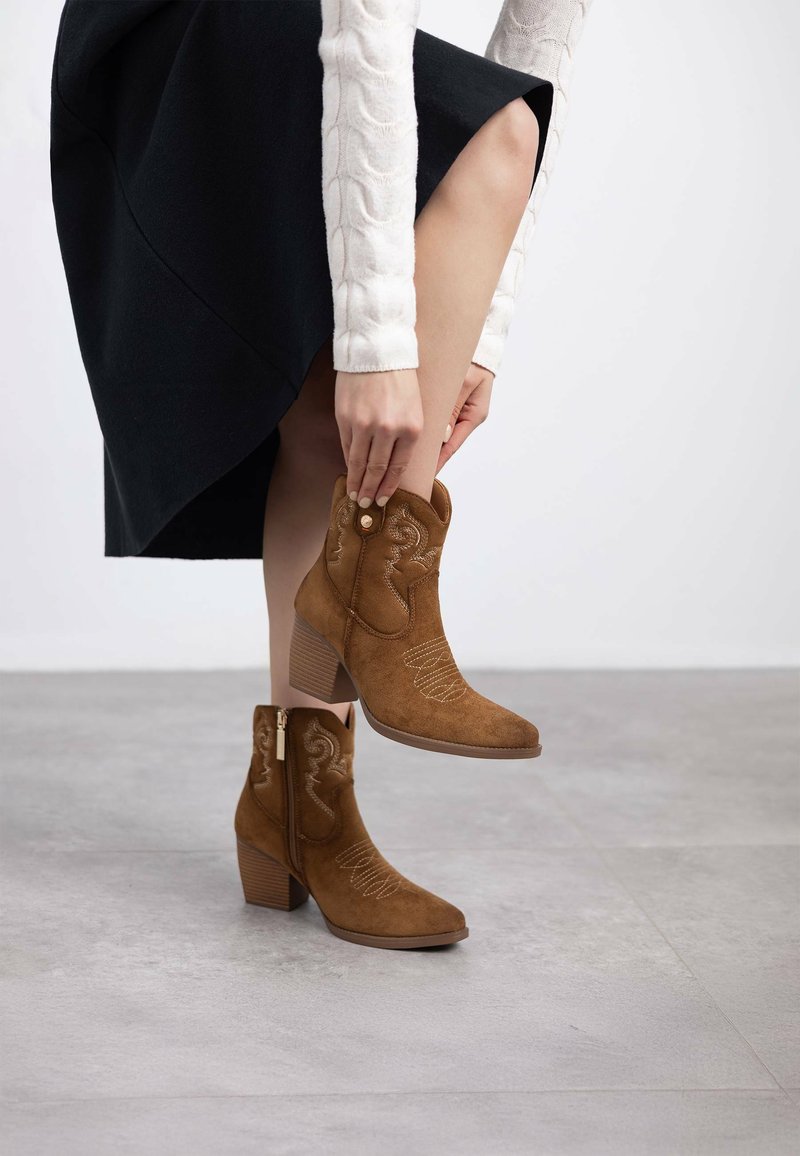 Botas de ante marrón hasta el tobillo con punta afilada, detalles de costura y un tacón cuadrado. Combinadas con una falda negra y un suéter de punto de cable color crema.