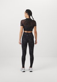 Top corto de malla negra con mangas cortas y leggings negros de cintura alta que presentan un patrón de camuflaje oscuro texturizado. Zapatillas deportivas blancas.