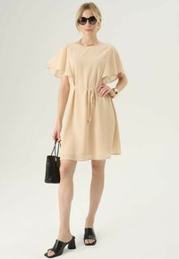 Abito corto beige con maniche a sbuffo e vita regolabile con cordino. Abbinato a mule nere con tacco bloccato e borsa nera dalla texture.