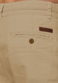 Khaki shorts made of soft fabric, featuring a rear pocket with a leather label and a button detail. Smooth texture and neutral color.