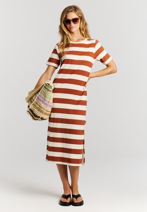 Vrouw die een bruin-wit gestreepte midijurk draagt, zwarte sandalen en een zonnebril, met een gevlochten meerkleurige tas in haar hand, staande tegen een effen achtergrond.