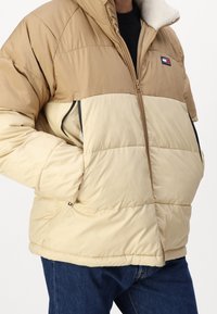 Puffer jacket featuring a tan and cream color block design, zip pockets, and a soft, textured hood. Material appears lightweight and insulated.
