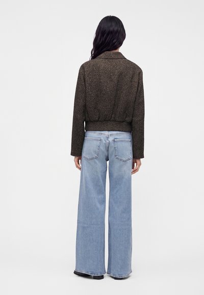 Femme aux longs cheveux foncés portant une veste marron texturée et un jean large bleu clair, debout de dos sur un fond uni.