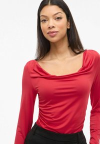 Czerwona bluzka z długim rękawem o kwadratowym dekolcie i marszczeniami. Gładki, elastyczny materiał. Połączona z czarnymi spodniami o wysokim stanie.