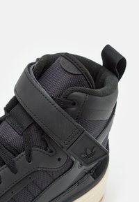 Hög sneaker med svart syntetiskt överdel och meshinslag, justerbar rem och texturerade detaljer. Gummisula i crèmefärg.