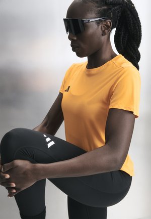 Femme portant des lunettes de soleil noires, un t-shirt orange à manches courtes et des leggings noirs, étirant une jambe en tenant son genou, sur un fond clair.