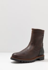 Ruskea nahkainen nilkkuri, jossa on pyöreä varvas, teksturoitu päällinen, ommellut yksityiskohdat ja kestävä musta kumipohja.