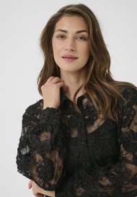 Blusa in pizzo nero con ricami floreali, chiusura con bottoni e polsini aderenti. Tessuto traslucido con dettagli testurizzati in tutta la superficie.