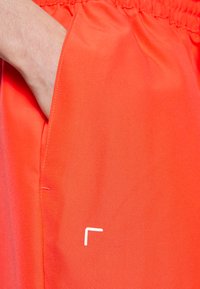 Handen delvis inne i sidofickan på ljusorangea shorts med elastisk midja, med en liten vit logotyp nära fickan.