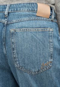 Ανοιχτό μπλε τζιν παντελόνι με πίσω τσέπη που διαθέτει ραμμένες λεπτομέρειες και μπεζ δερμάτινο έμπλαστρο κοντά στη μέση.