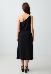 Schwarzes, asymmetrisches Kleid mit einem Schulterträger; knielang, figurbetontes Oberteil, fließender Rock. Getragen mit schwarz-gestrickten Blockabsatzsandalen.