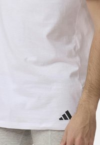 Biała bawełniana koszulka z okrągłym dekoltem, z czarnym logo Adidas na dolnym lewym boku i gładką strukturą materiału.