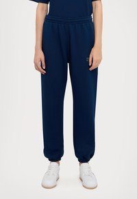 Navyblauwe joggingbroek van zacht materiaal met een elastische tailleband en boorden. Voorzien van een klein logo aan de voorkant. Gepaarde met witte sneakers.