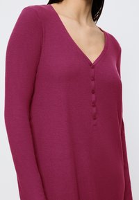 Donna che indossa una maglia a maniche lunghe di colore rosa intenso con scollo a V e cinque bottoni sul petto.