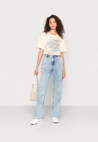 T-shirt gráfica bege com uma estampa circular, jeans de cintura alta azul clara, ténis brancos e uma mala de ombro bege clara.